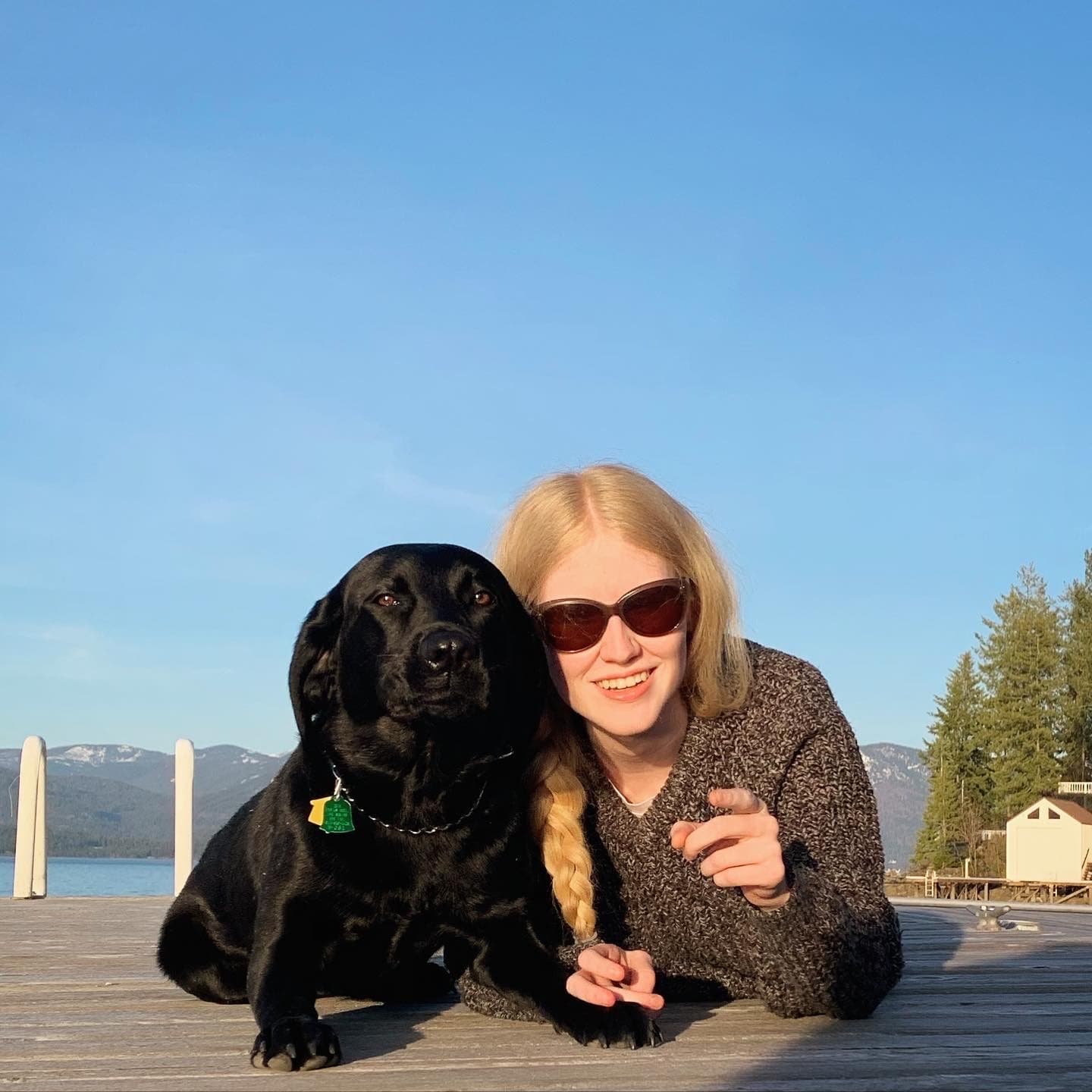 Freida, a black Lab, lies next to Katherine on a dock overlooking a large lake and an array of mountains.