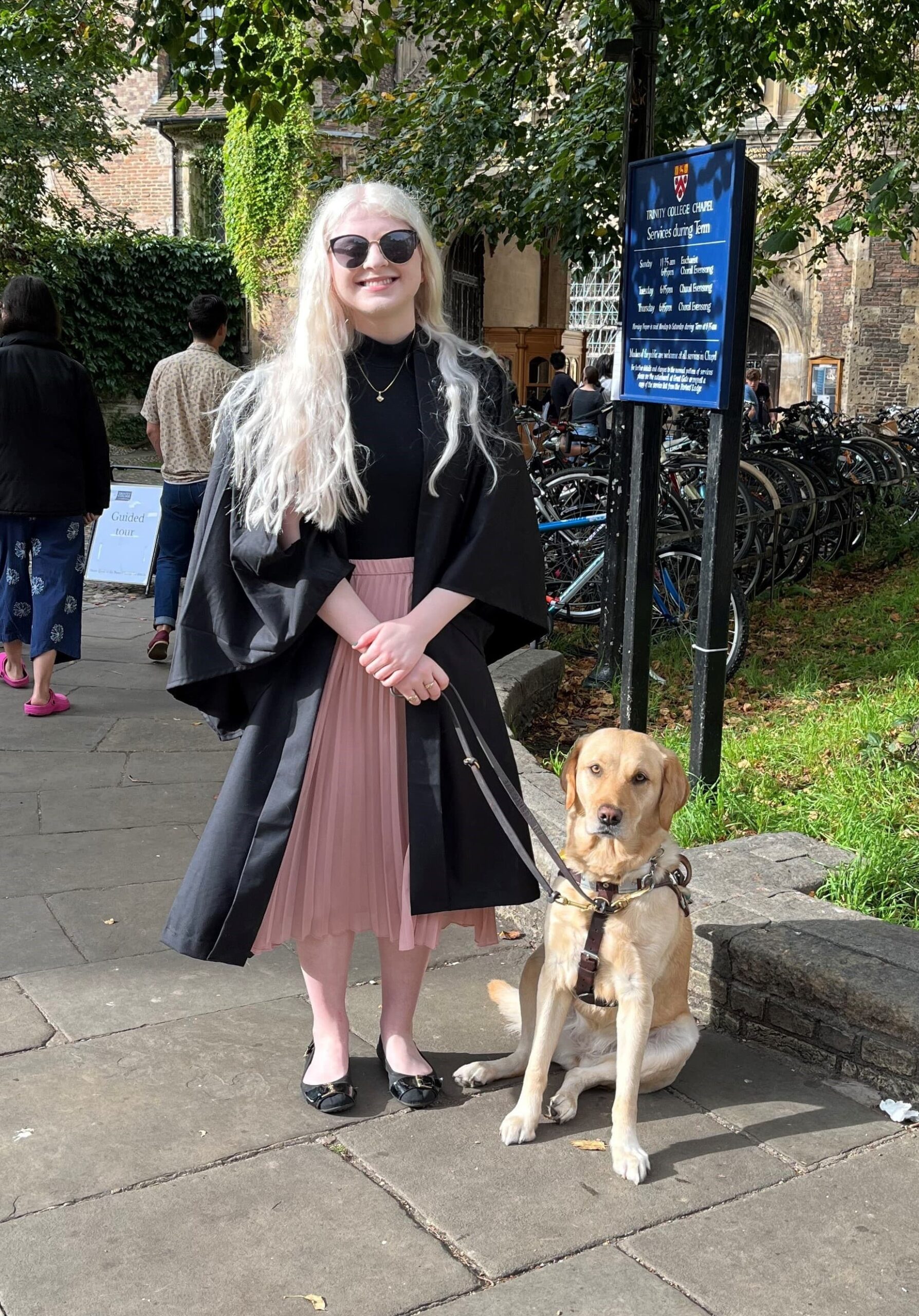 Sarah stands with her yellow Lab/golden cross Seeing Eye dog, Elana, sitting by her side in front of the Trinity College Chapel at the University of Cambridge.