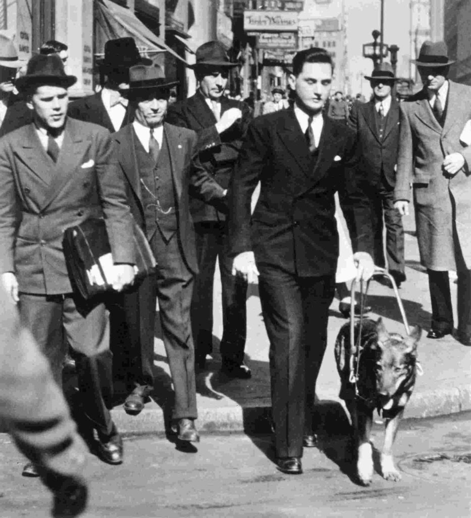 Black and white archival image with Morris Frank and Buddy stepping off a curb before a crowd of onlookers.