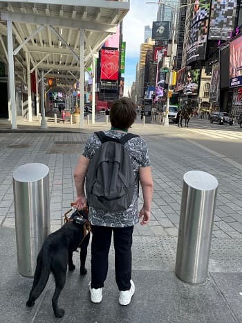 A Seeing Eye dog handler stands at the down curb of a sidewalk in New York City with their black Lab Seeing Eye dog.