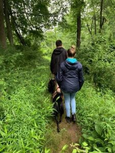 Two students with black Labs are hiking on a forest trail while in class at The Seeing Eye. A young man is in front and a young woman follows.