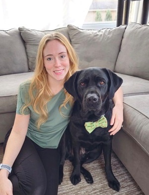 Alyssa sits on the ground before a sectional with her arm wrapped around her first Seeing Eye dog, a black Lab/golden retriever cross named Autumn, who wears a green bow on her collar.