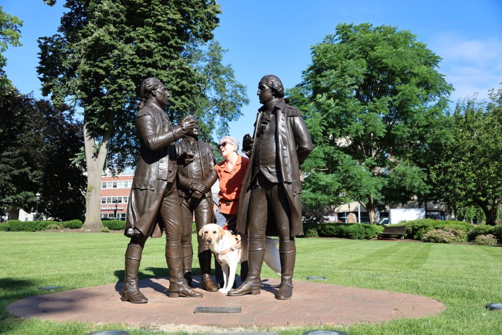 A Seeing Eye student, guided by a yellow Labrador retriever, stands among three brass statues of George Washington, Alexander Hamilton, and the Marquis de Lafayette on the Morristown Green.