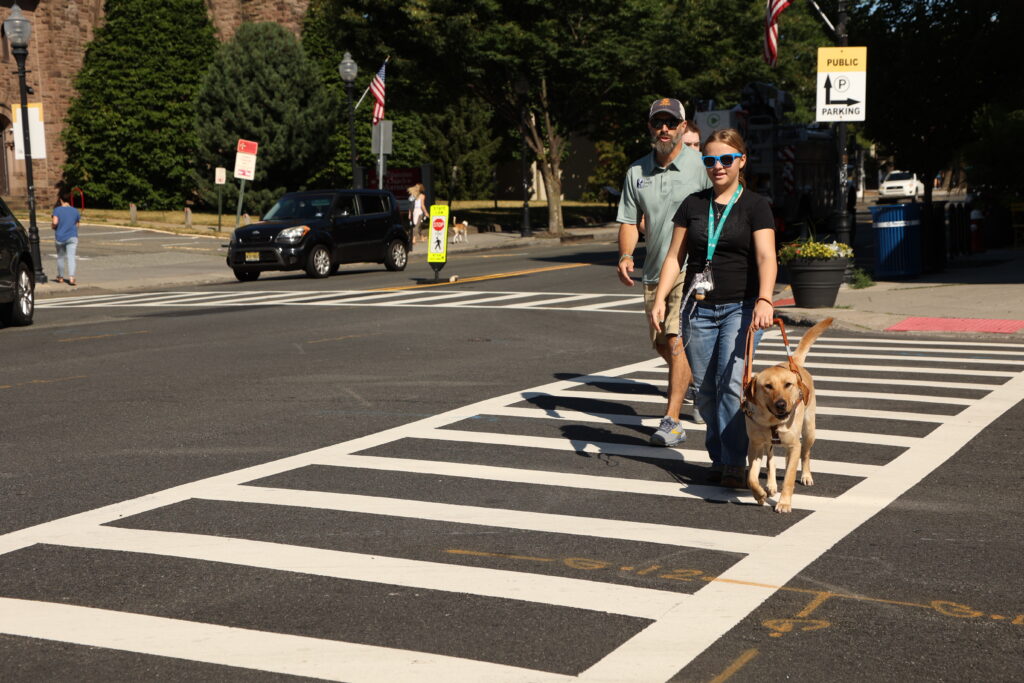 A Seeing Eye instructor works with a student in class as she is guided across a street by a yellow Labrador retriever.