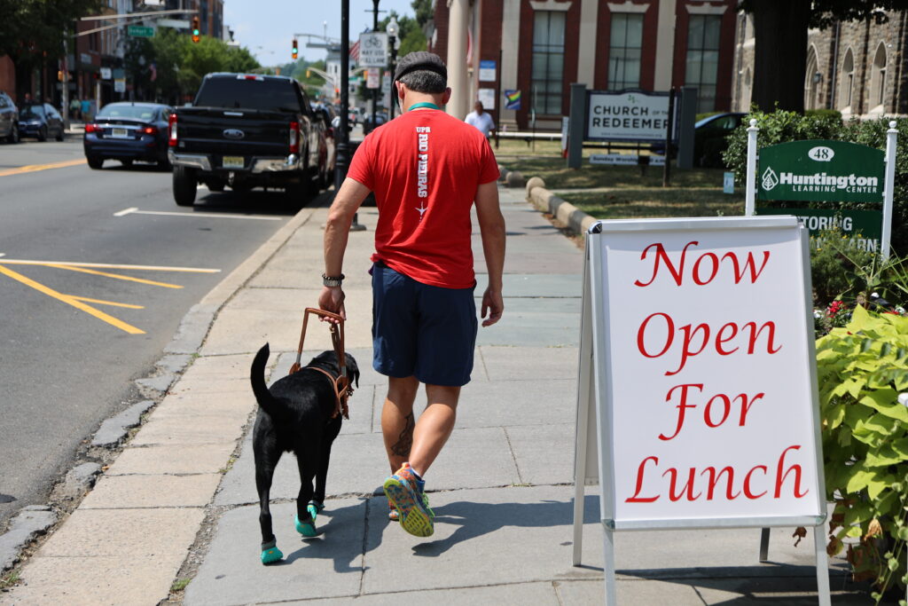 A Seeing Eye graduate is led by a black Labrador retriever on a sidewalk past a sign reading 'Now Open for Lunch'.