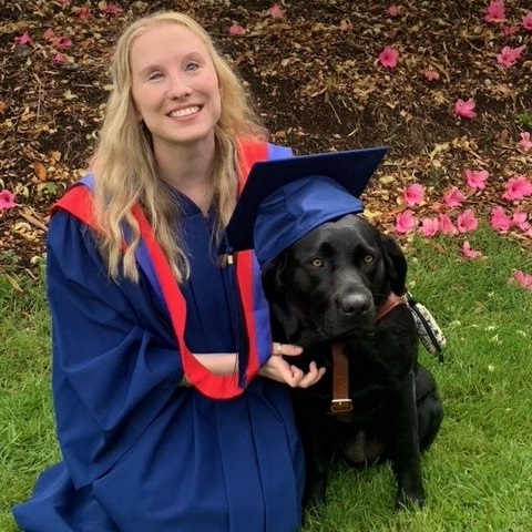 A Seeing Eye graduate, wearing a graduation gown, hugs her Seeing Eye dog, a black Labrador retriever wearing a graduation cap.