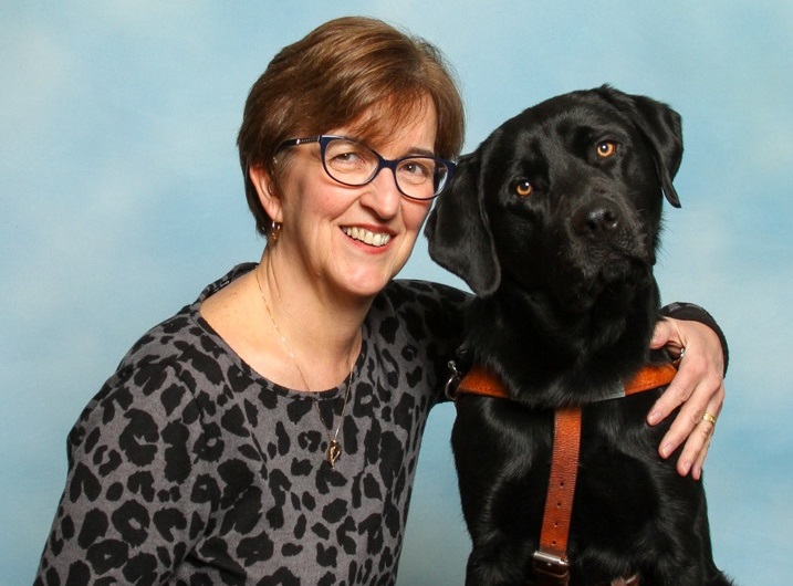 A smiling woman with her arm around a black Labrador retriever.