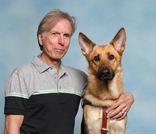 A man with his arm around a German shepherd, in harness.