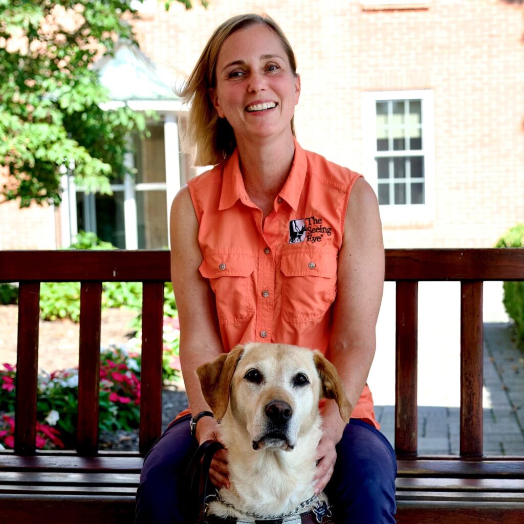 A woman with straight blonde hair sits on a wood bench with her yellow Lab/golden cross in harness and seated between her knees. Both look at the camera, the dog with her ears perked alertly, the woman smiling.