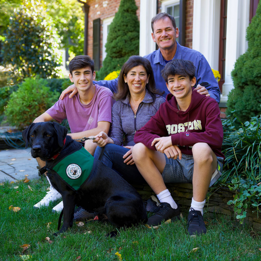 A puppy raising family posing together outside of their home with their black Lab Seeing Eye puppy, who wears a Seeing Eye puppy raising program vest.