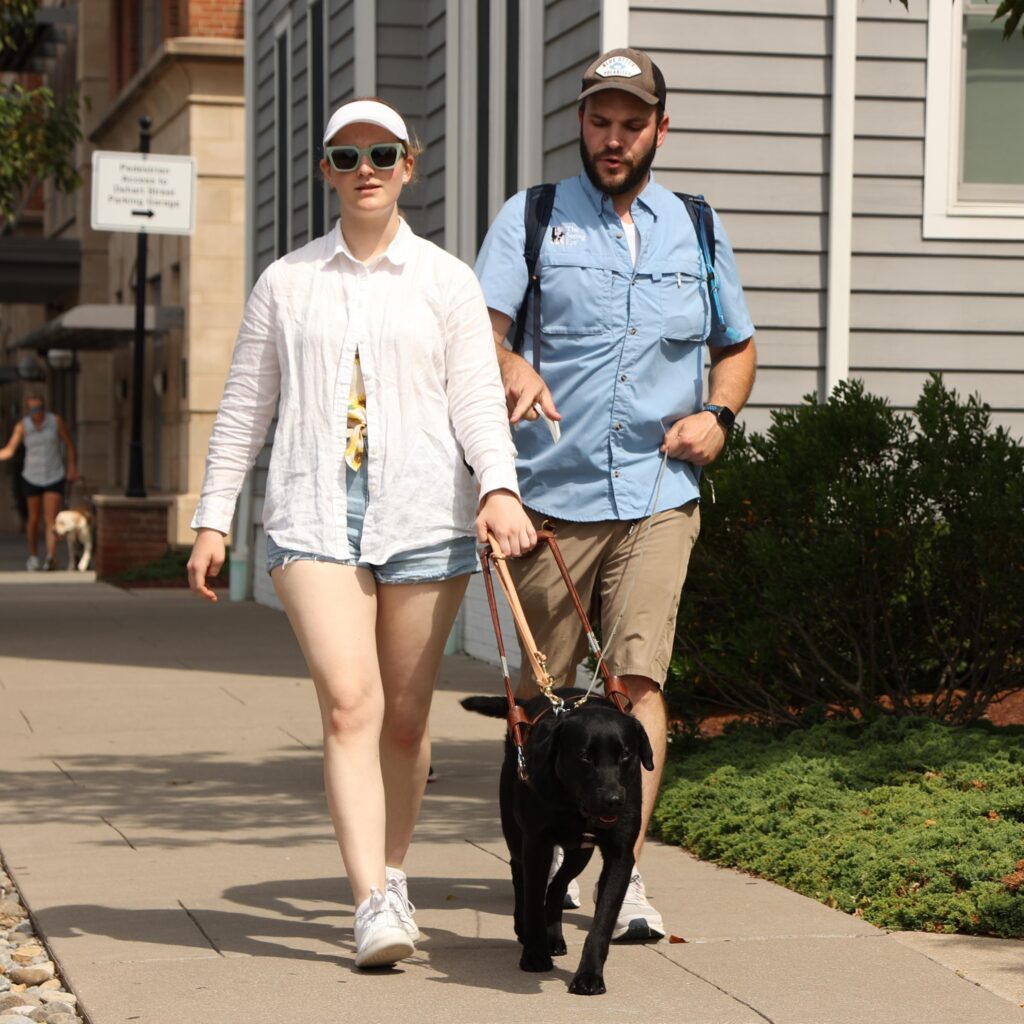 A woman wearing sunglasses is guided by a black Lab down a sidewalk with an instructor walking beside her.