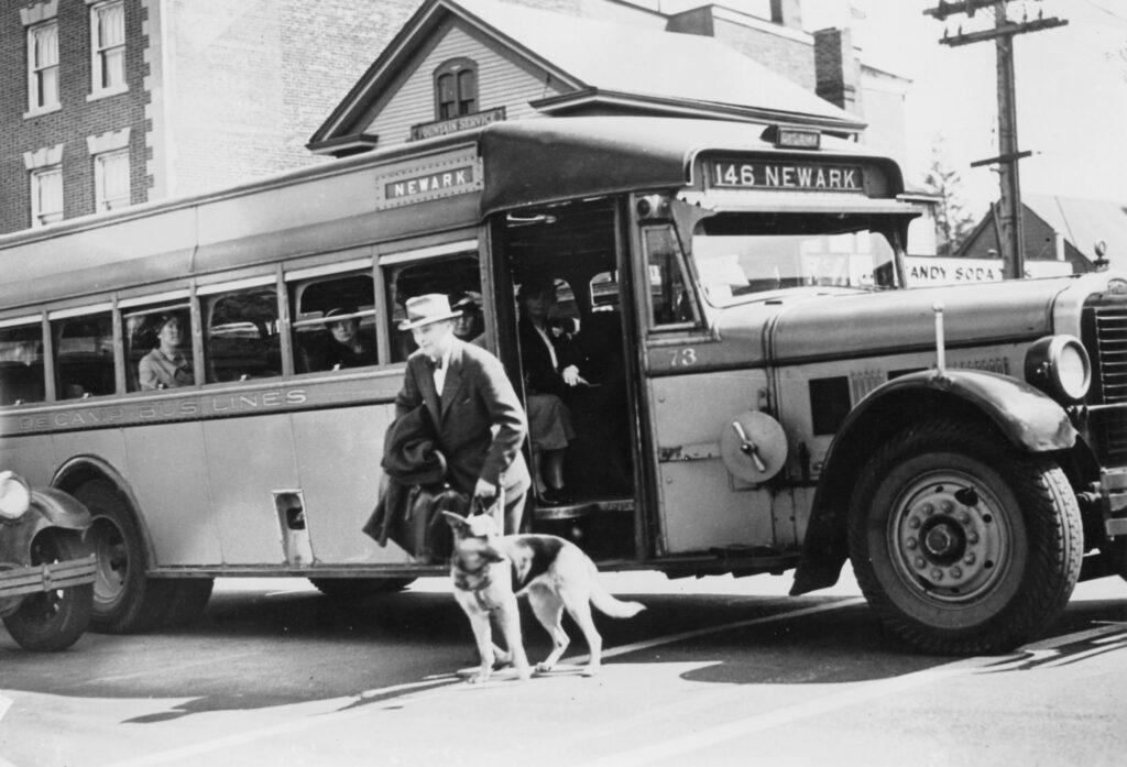 A historic black and white photo of a man being guided by his Seeing Eye dog off a city bus.