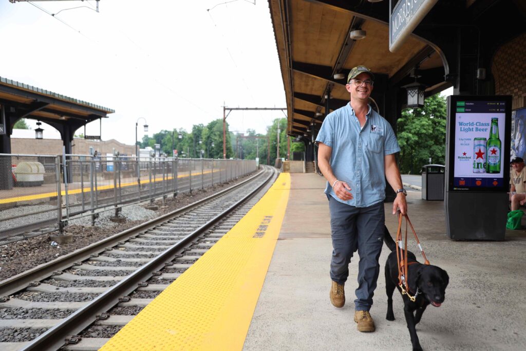 A male guide dog mobility instructor is guided along a train platform by a black Lab.