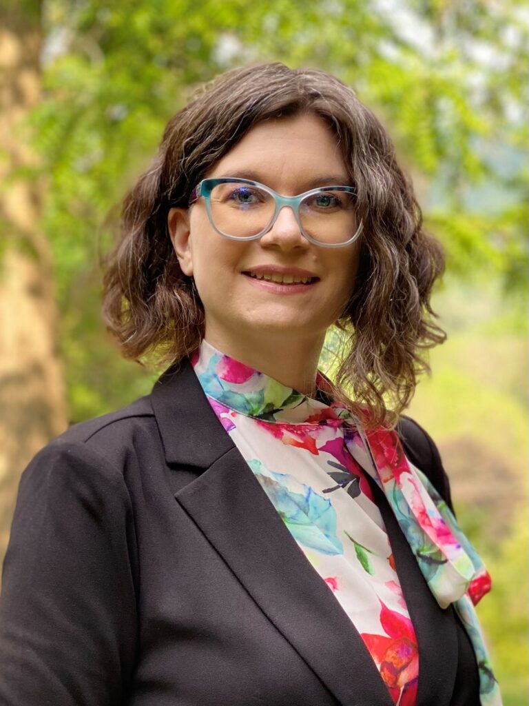 A woman with curly brown hair smiles. She is wearing blue glasses, and a black suit with a colorful blouse.