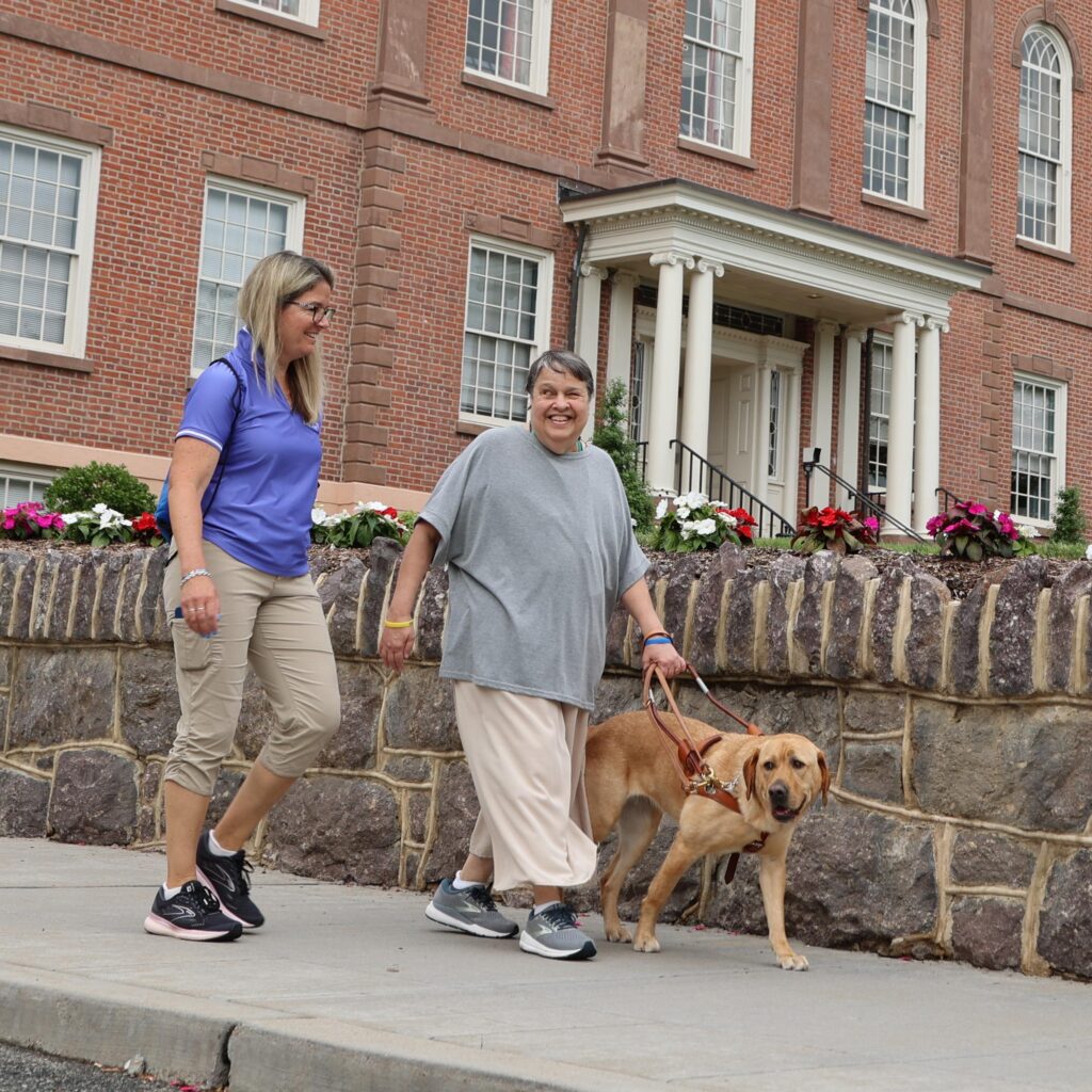 A female instructor walks down a sidewalk beside a female student with her guide dog.