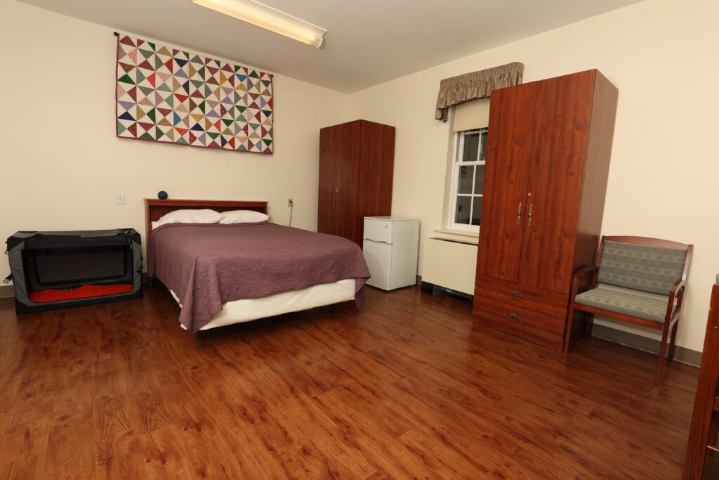 One of the student rooms on The Seeing Eye campus with a window and a hardwood floor. There is a bed, two wardrobes, a mini-fridge, a soft crate, an Echo Dot, a chair, and the corner of a desk.