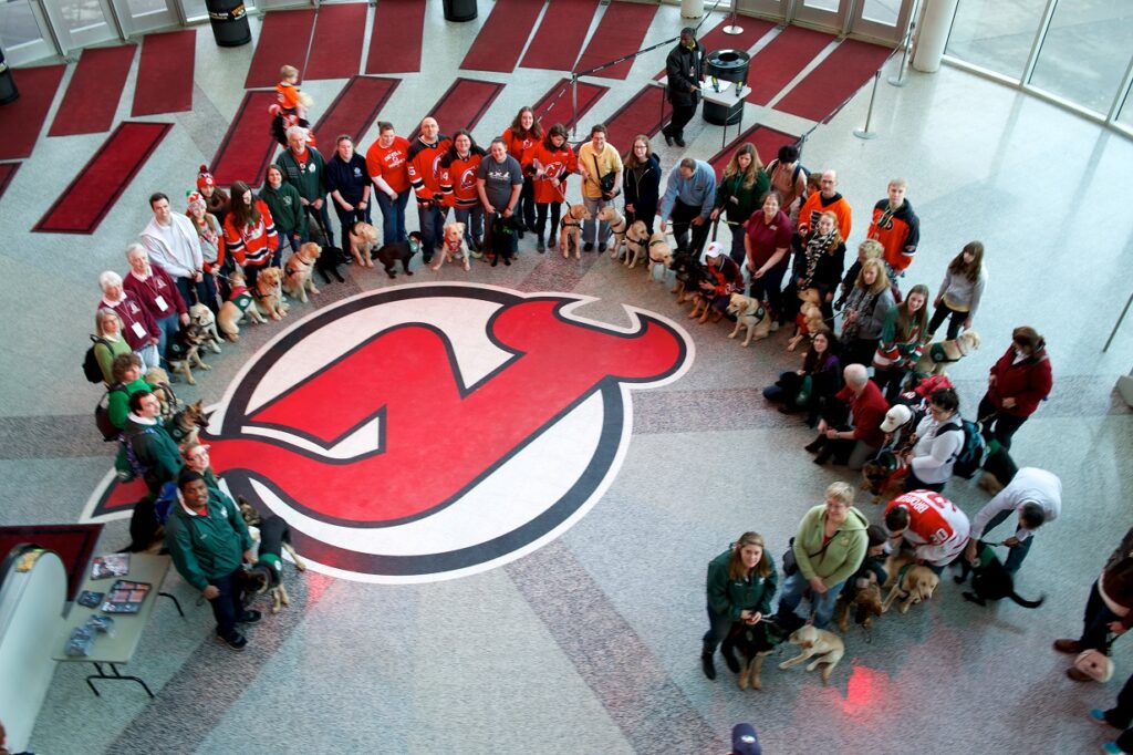 A birds-eye-view of a large group of puppy raisers with their Seeing Eye puppies circling the NJ Devils logo.