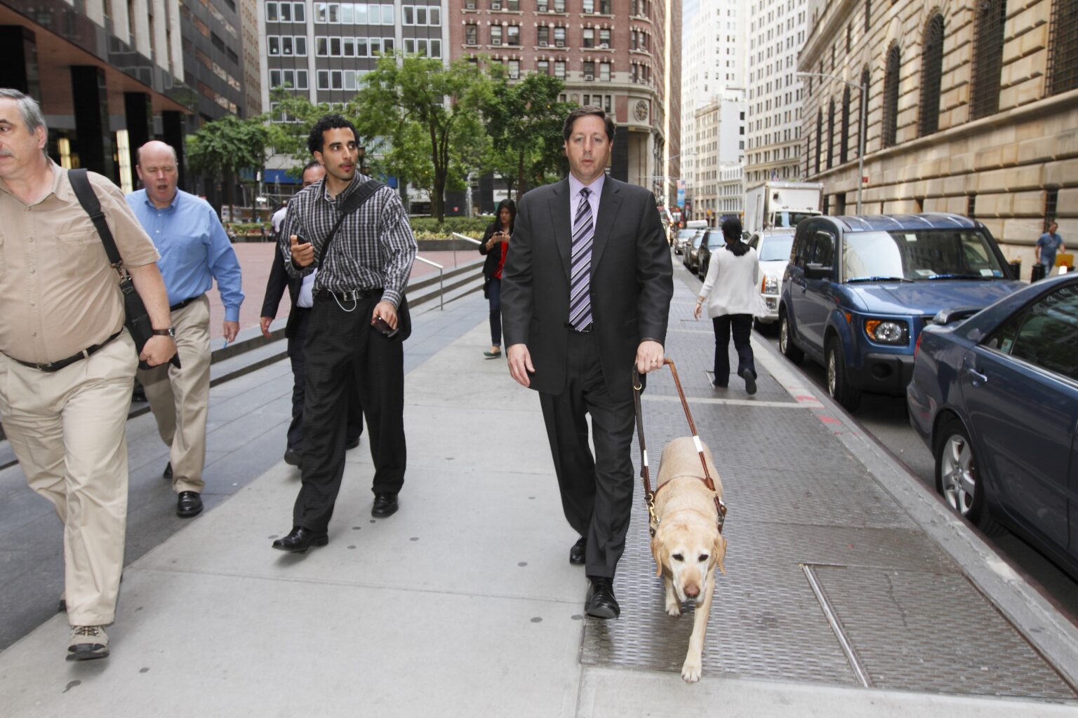 A Seeing Eye graduate, wearing a suit, being guided by a yellow Labrador/golden retriever cross in New York City.