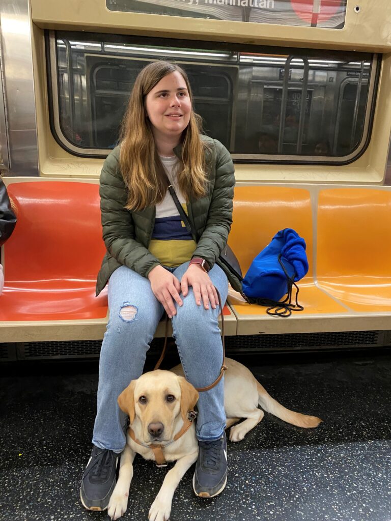 Seeing Eye graduate Kylie smiles as she sits on the New York City subway with Olivia, her two-year-old yellow Lab Seeing Eye dog tucked under the seat and between Kylie’s feet.