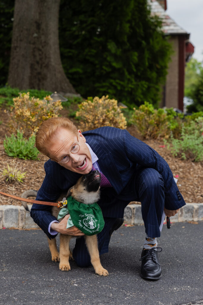 John Elliott smiles as he kneels to pet a German shepherd wearing a Seeing Eye puppy raising program bandana. The puppy leans her head up to give John a kiss.