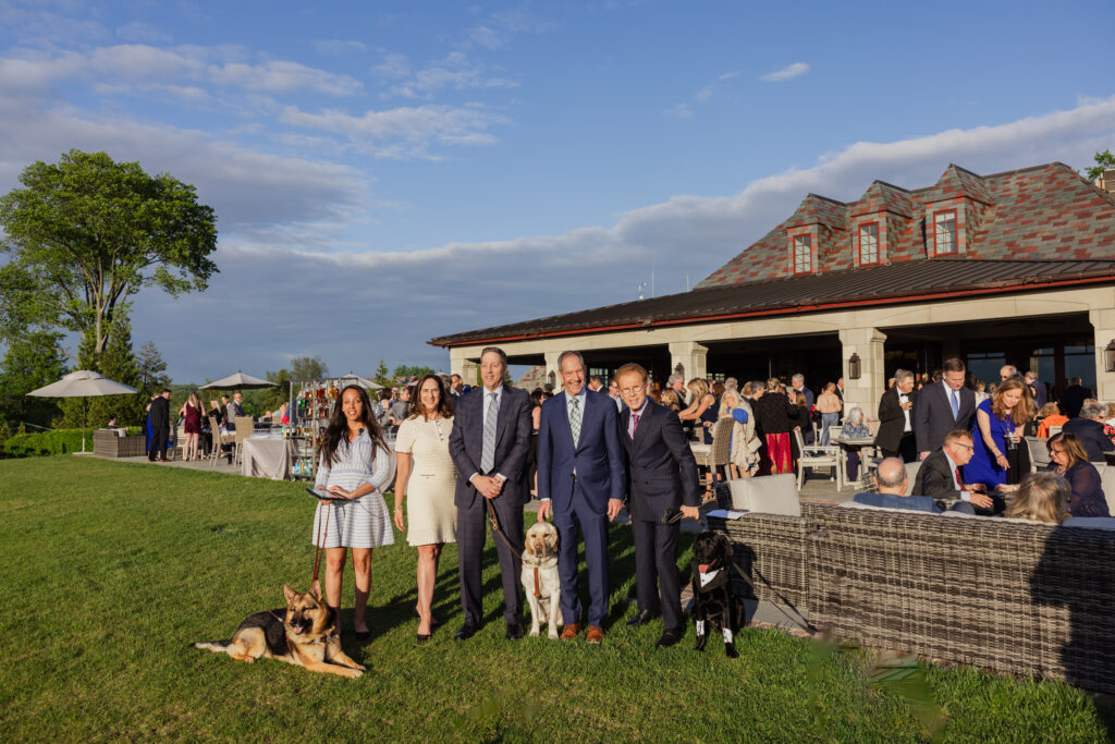 Haben Girma, Karen Leies, Cliff Aaron, Mike Mittelman, and John Elliott pose together as guests mingle during cocktail hour on the Mountain Ridge Country Club patio behind them. Haben is with her German shepherd Seeing Eye dog, Cliff is with his yellow Lab Seeing Eye dog, and John is with a black Lab Seeing Eye puppy, dressed in a suit, patterned bandana, and cufflinks.
