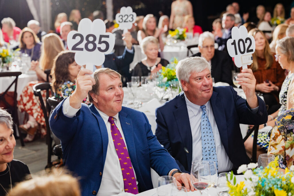 Two gentlemen smile as they lift their pawprint-shaped auction paddles at their table.