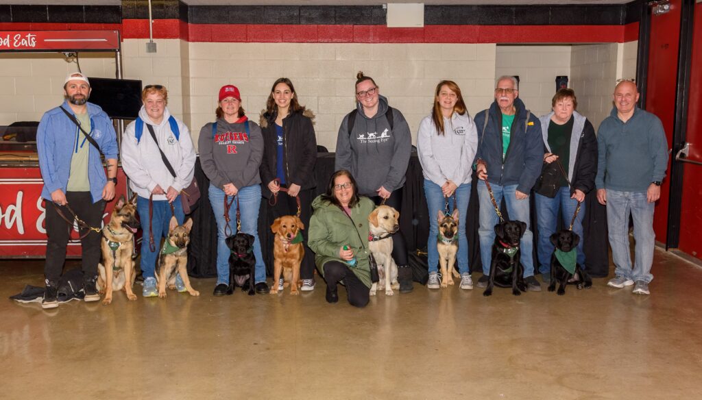 Ten Middlesex County puppy raisers line up with three German shepherds, three black Labs, a golden retriever, and a yellow Lab inside the Rutgers women’s basketball arena.