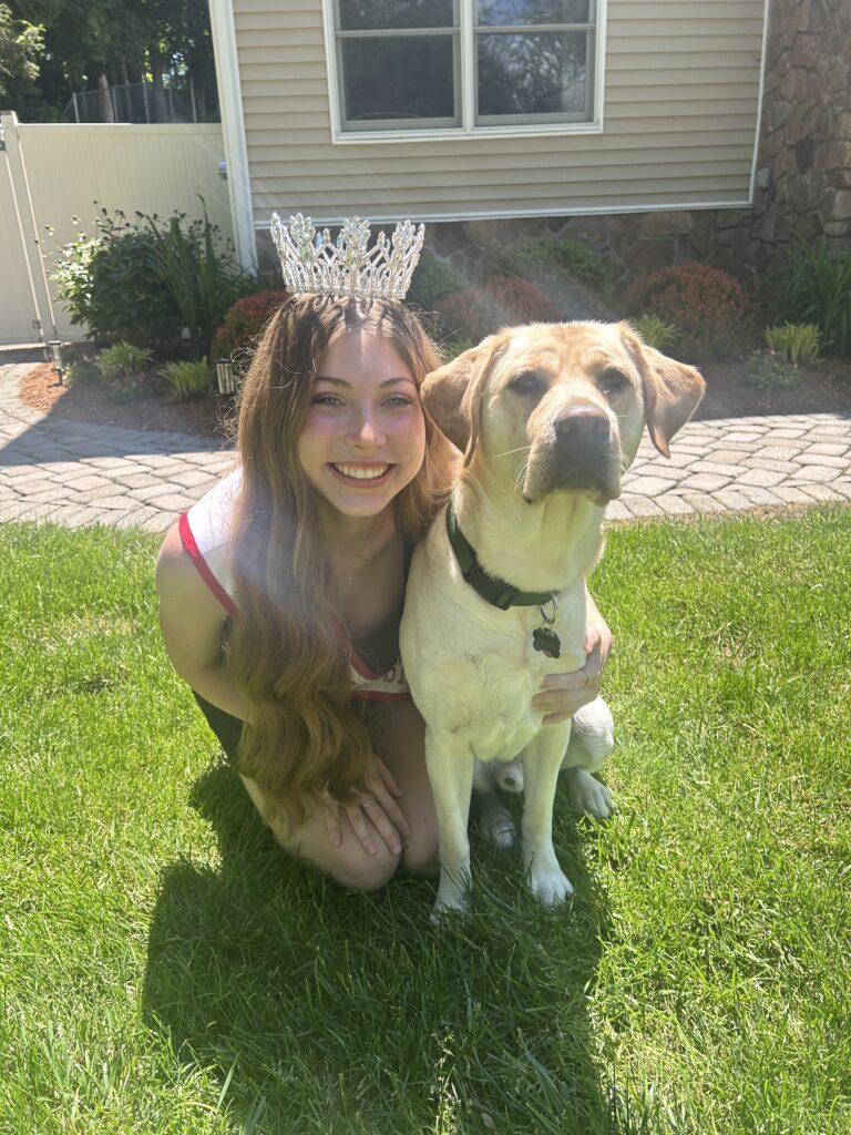 Makayla, wearing her tiara and sash for Miss Garden State Teen, kneels beside Zayde, a yellow Lab Seeing Eye puppy, in her backyard.