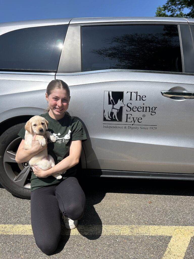 Makayla kneels beside a silver van with The Seeing Eye’s logo on the side of the door as she holds Katherine, a seven-week-old yellow Lab Seeing Eye puppy.