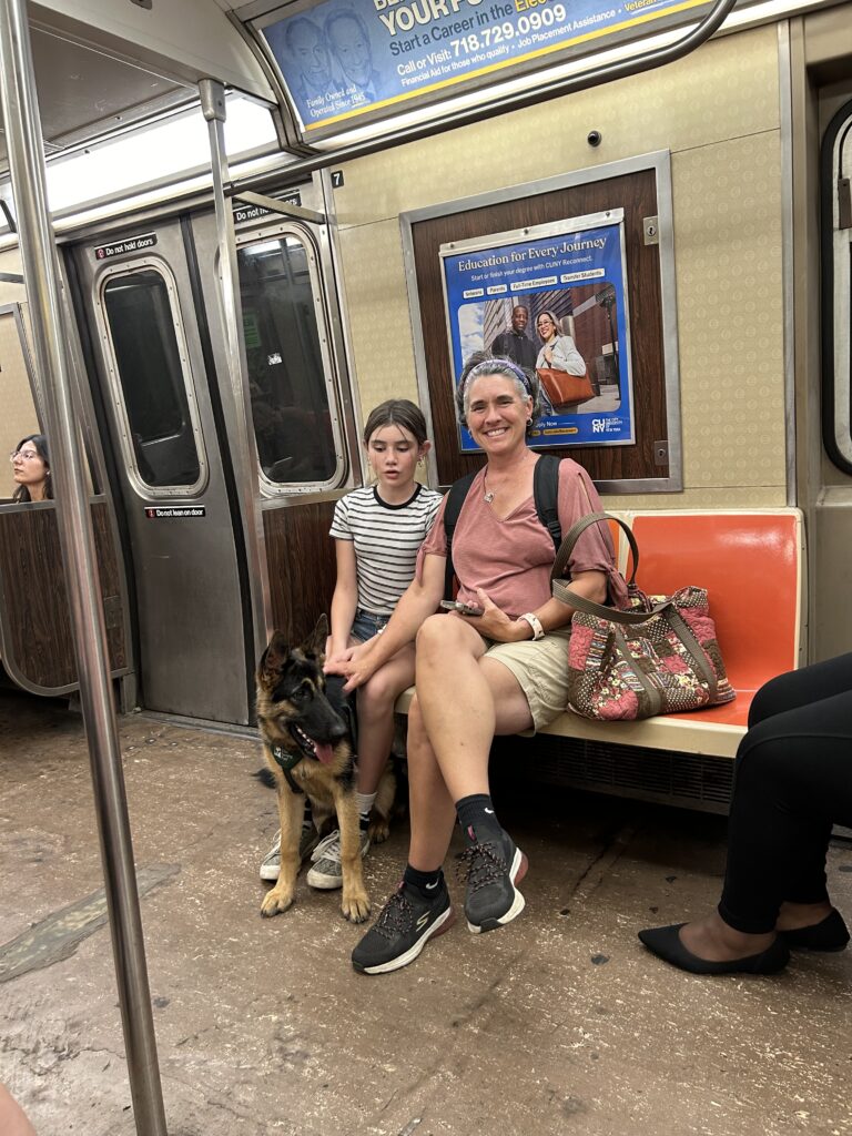 Melissa Sneider and her eleven-year-old daughter sit together on the New York City subway with Emme, a German shepherd wearing a Seeing Eye puppy raising program vest, sitting between her daughter’s feet.