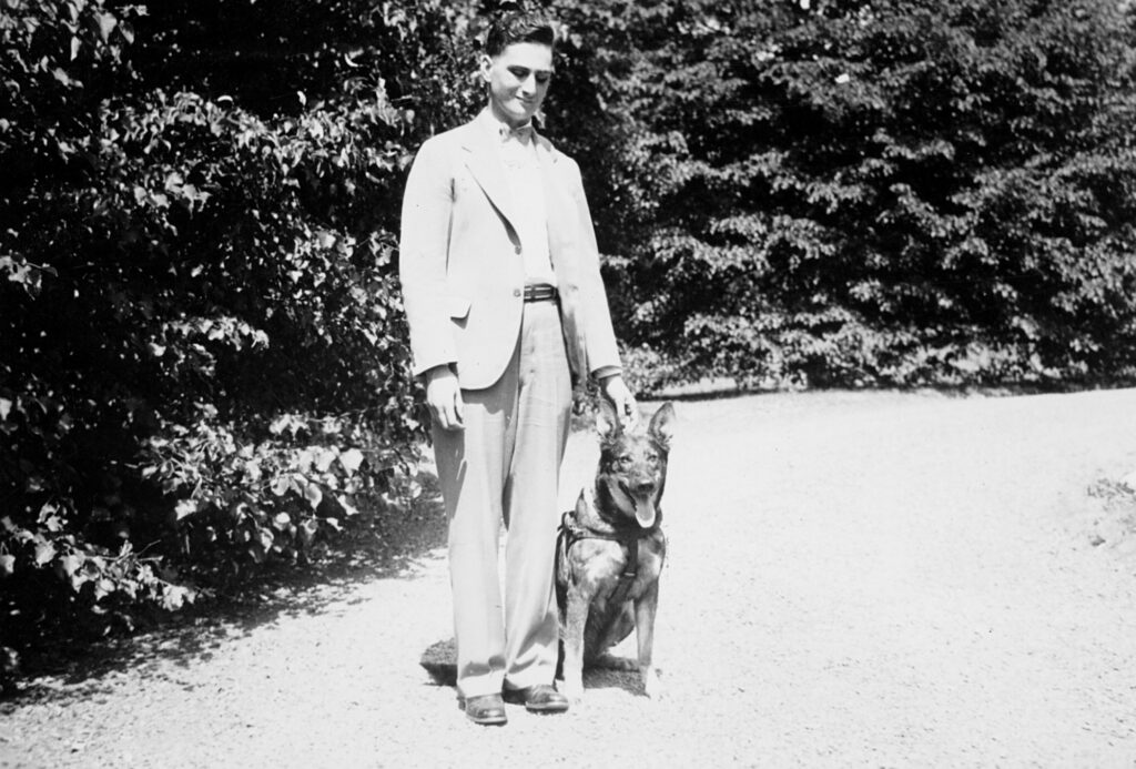 A black and white photo of a slightly smiling twenty-year-old Morris Frank standing with Buddy, his German shepherd Seeing Eye dog, sitting at his side. 