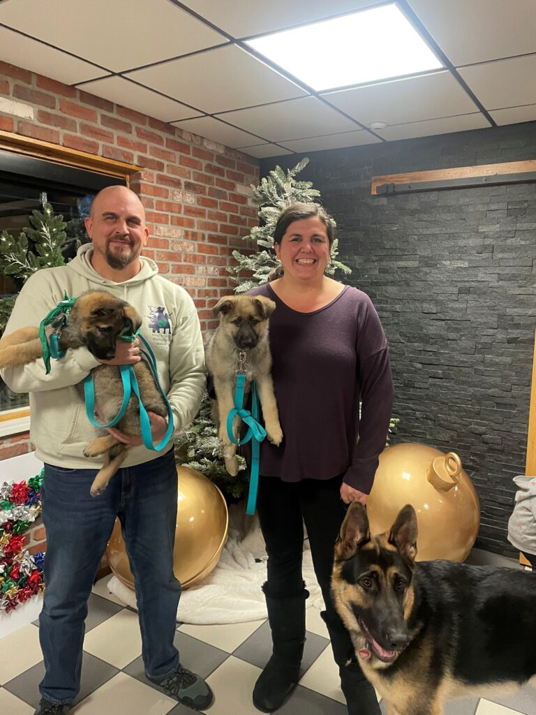 Scott and Laura pose together holding German shepherd puppies with an older German shepherd puppy standing beside Laura.