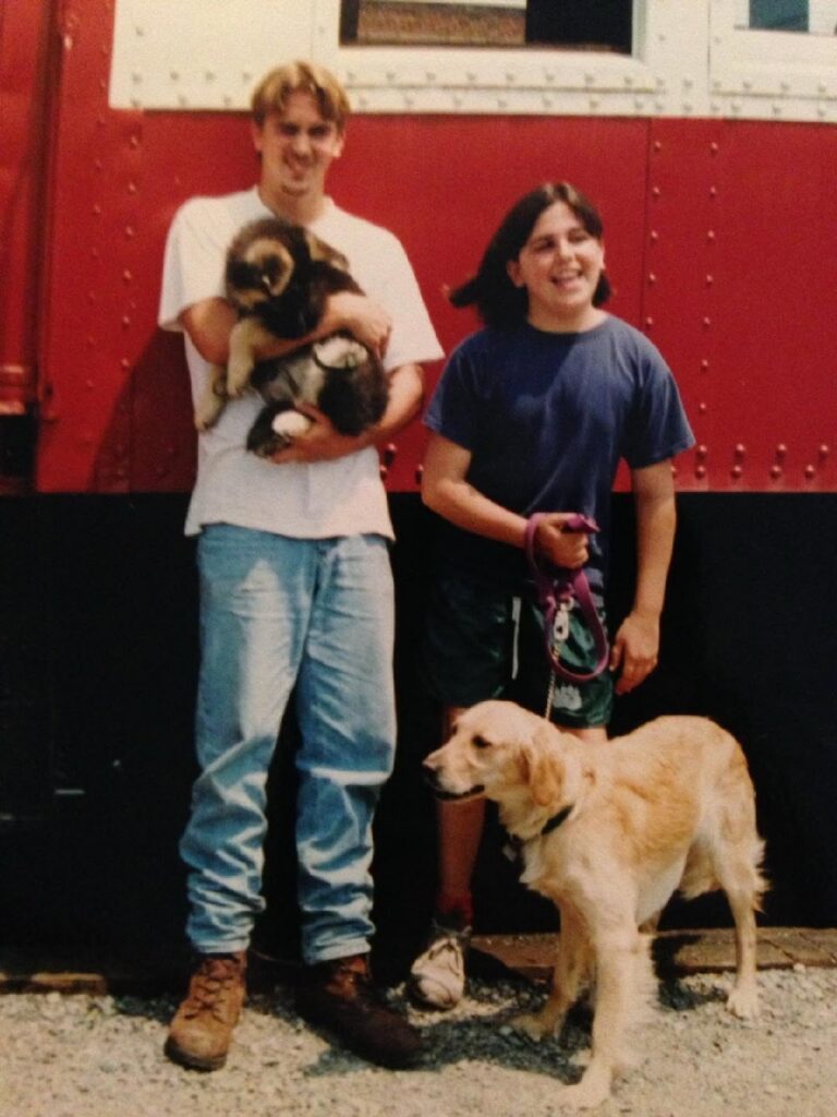 A throwback photo of Scott and Laura with the first puppies they raised. Scott holds a German shepherd puppy in his arms, and a golden retriever stands beside Laura.