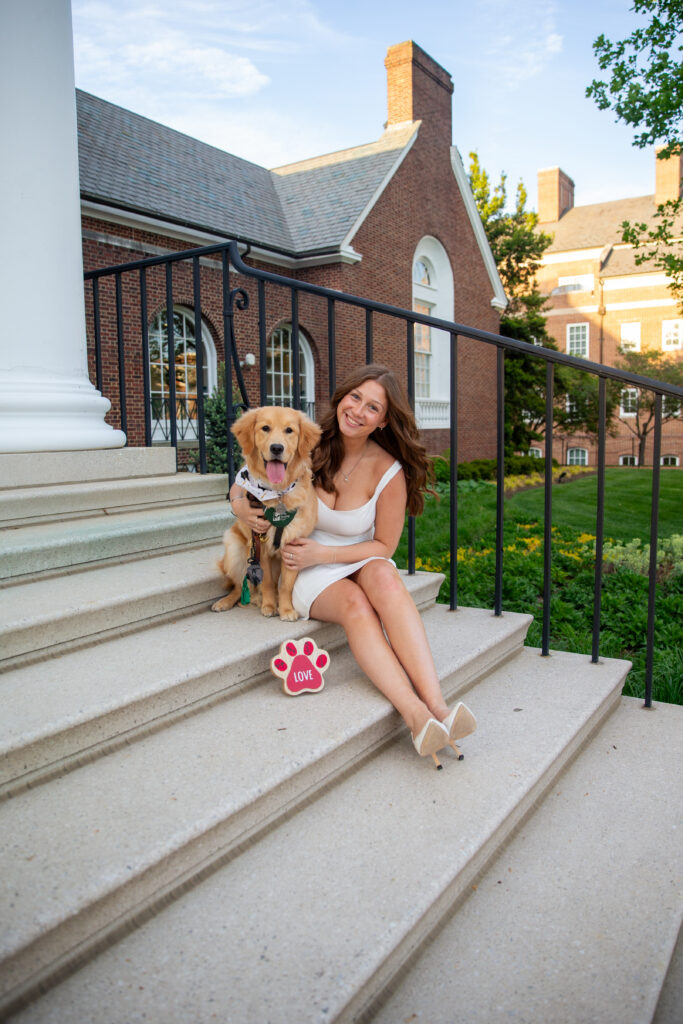 A posed graduation photo of Lillian Weingarten with Frances, the golden retriever Seeing Eye puppy she is currently raising, sitting on steps at the University of Delaware’s campus.