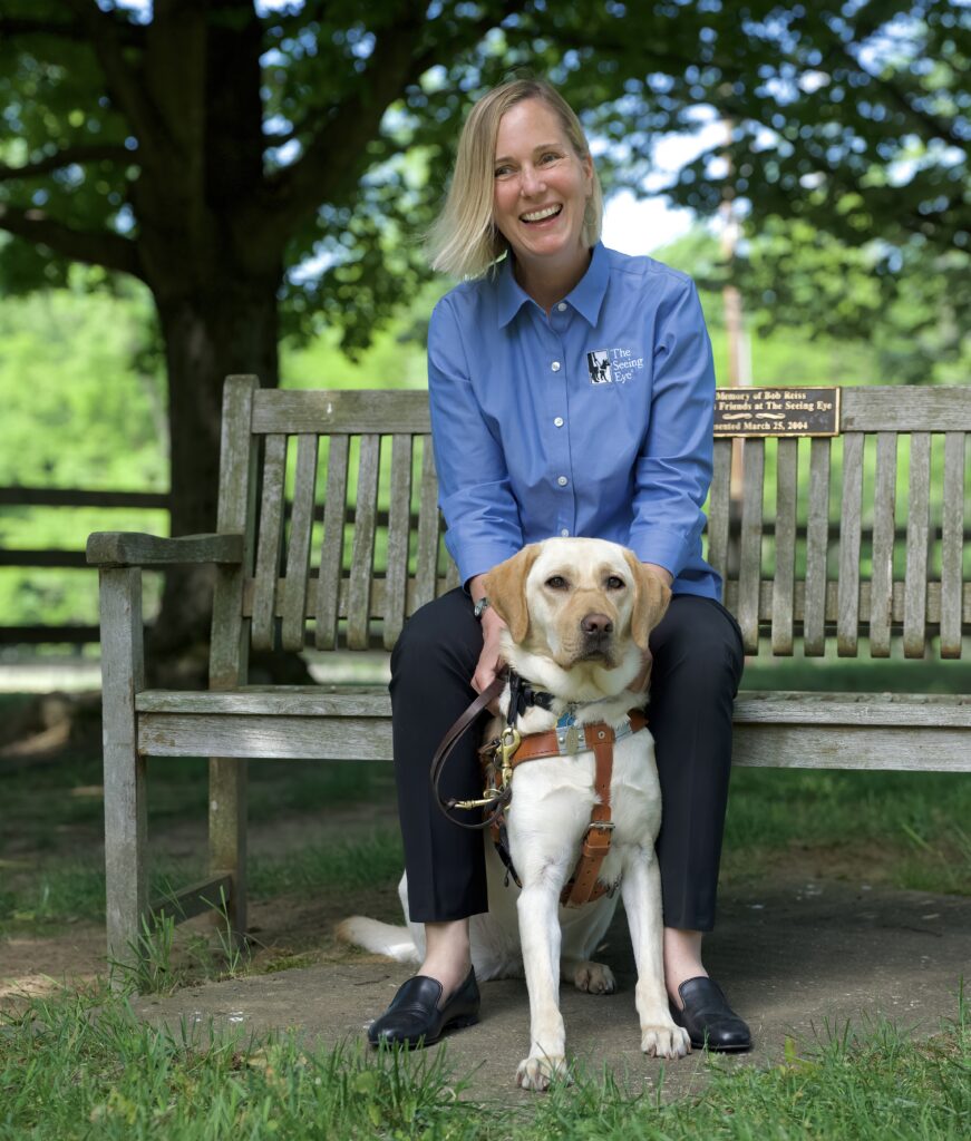 A woman with straight blonde hair sits on a wood bench with her yellow Labradpr retriever in harness and seated between her knees.